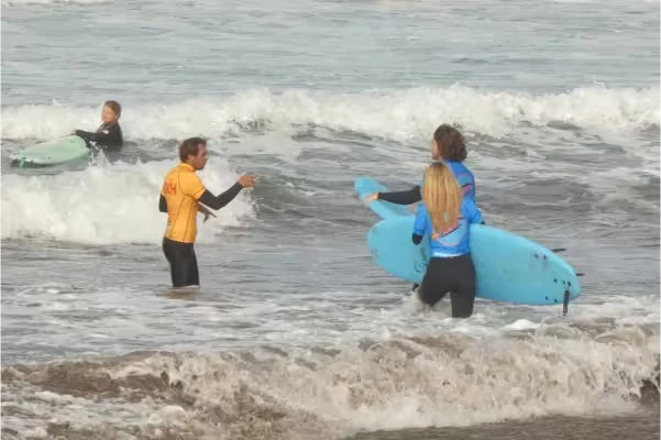 A private surf coach and two surfers entering the water with their surfboards as waves break around them.