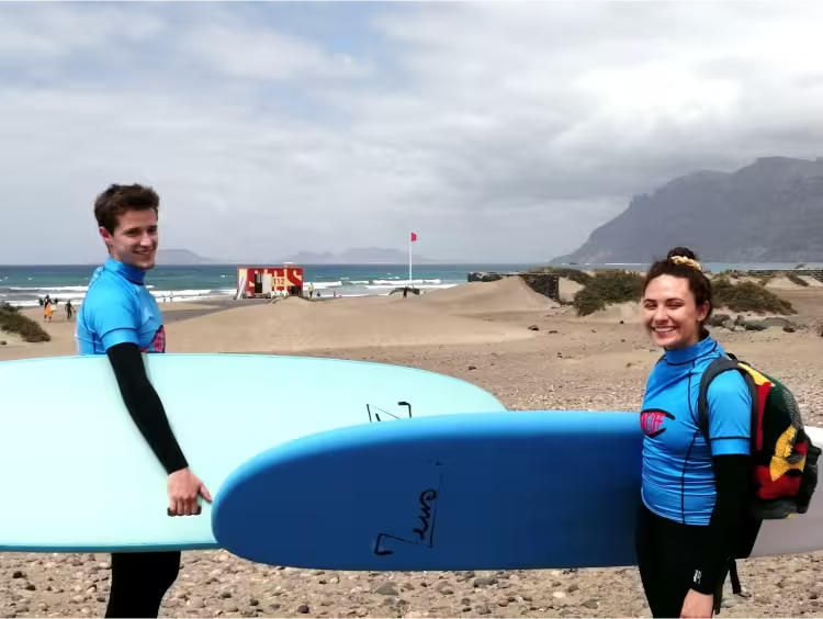 2 persons equiped with wetsuit, take-off surfschool lycra, and surfboards, carrying backpacks on their backs, smiling towards the camera in front of a beach panorama, ready to start their surflesson
