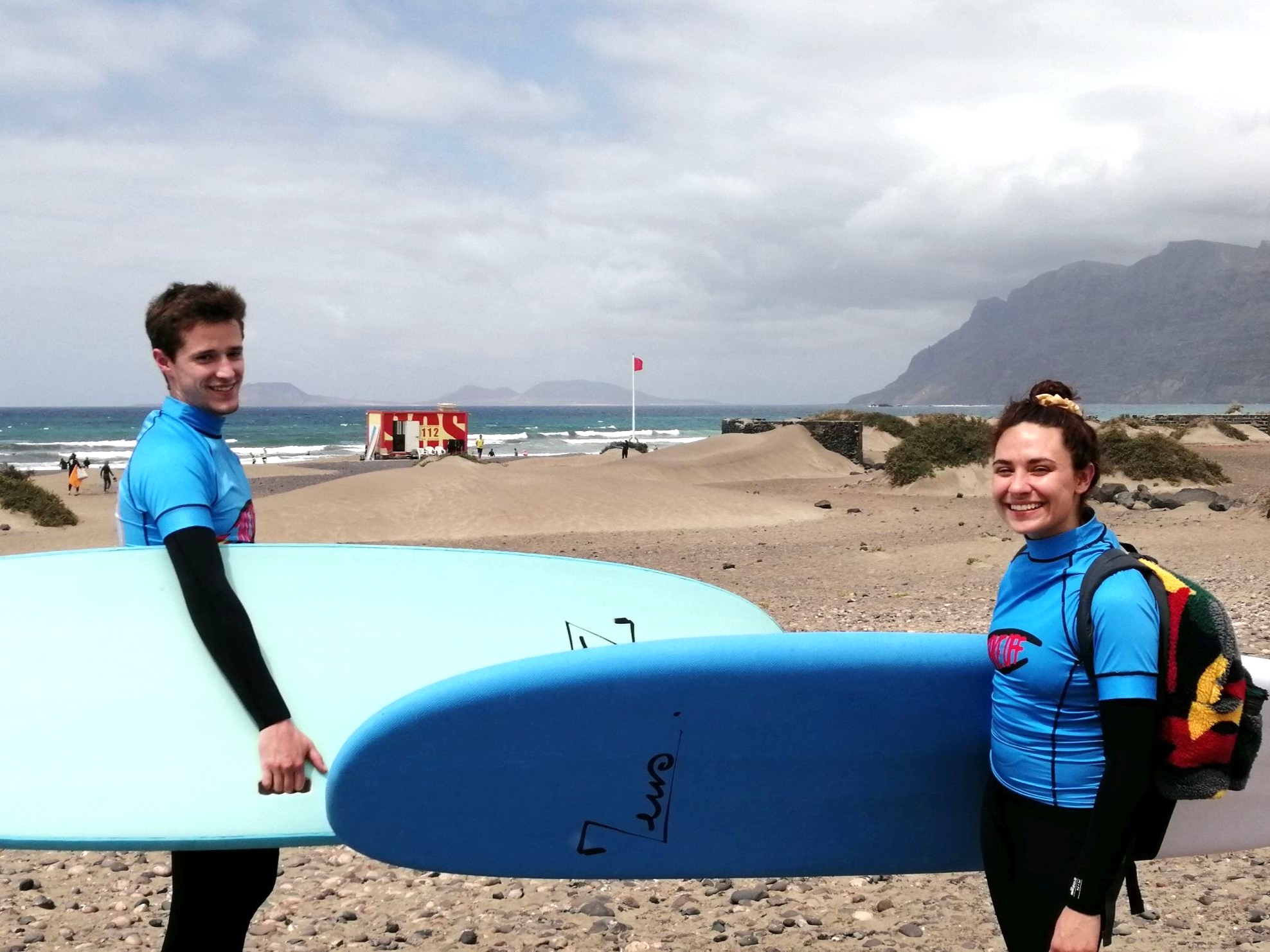 2 persons equiped with wetsuit, take-off surfschool lycra, and surfboards, carrying backpacks n their backs, smiling towards the camera in front of a beach panorama, ready to start their surflesson
